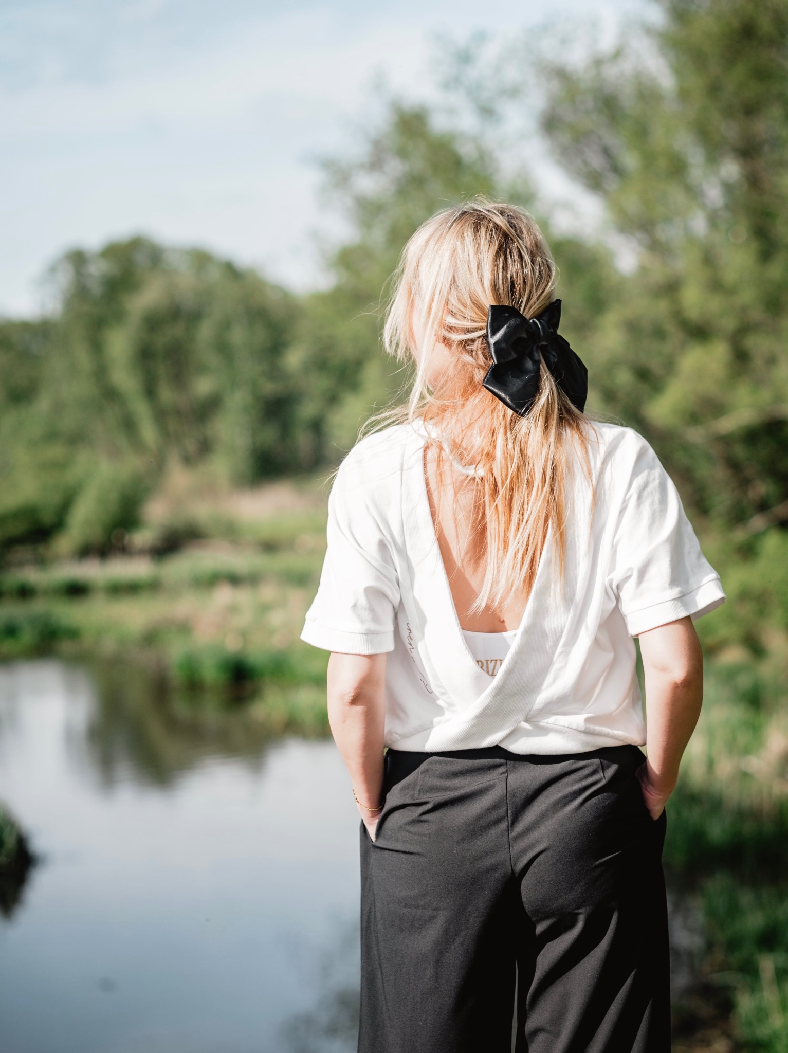 Kobieta, tyłem, z blond włosami upiętymi czarną kokardą, ubrana w białą koszulkę z odkrytymi plecami i czarne spodnie, stoi nad brzegiem rzeki, patrząc w dal.