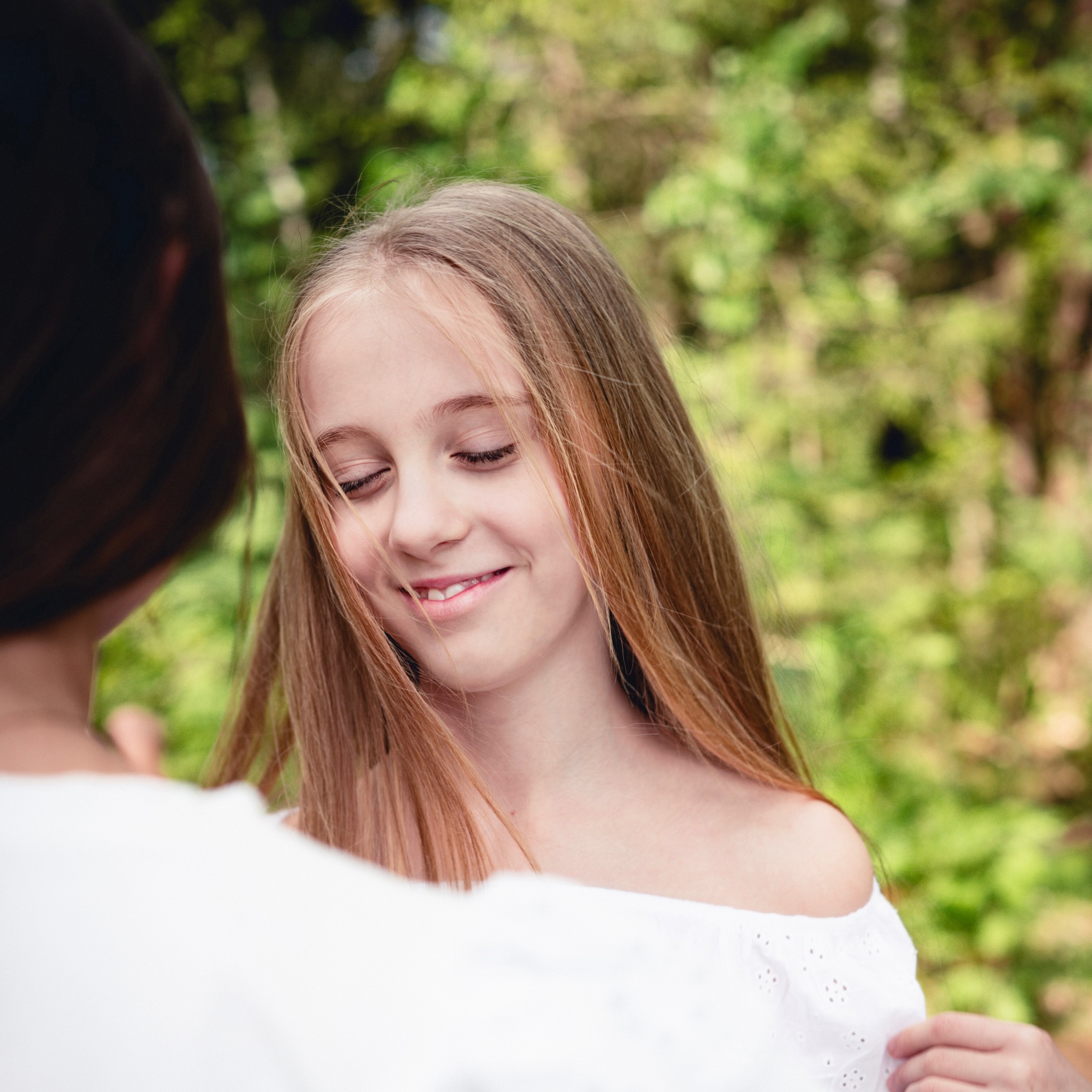 Portret uśmiechniętej nastolatki o długich blond włosach, w białej bluzce z odkrytymi ramionami, na tle zieleni.