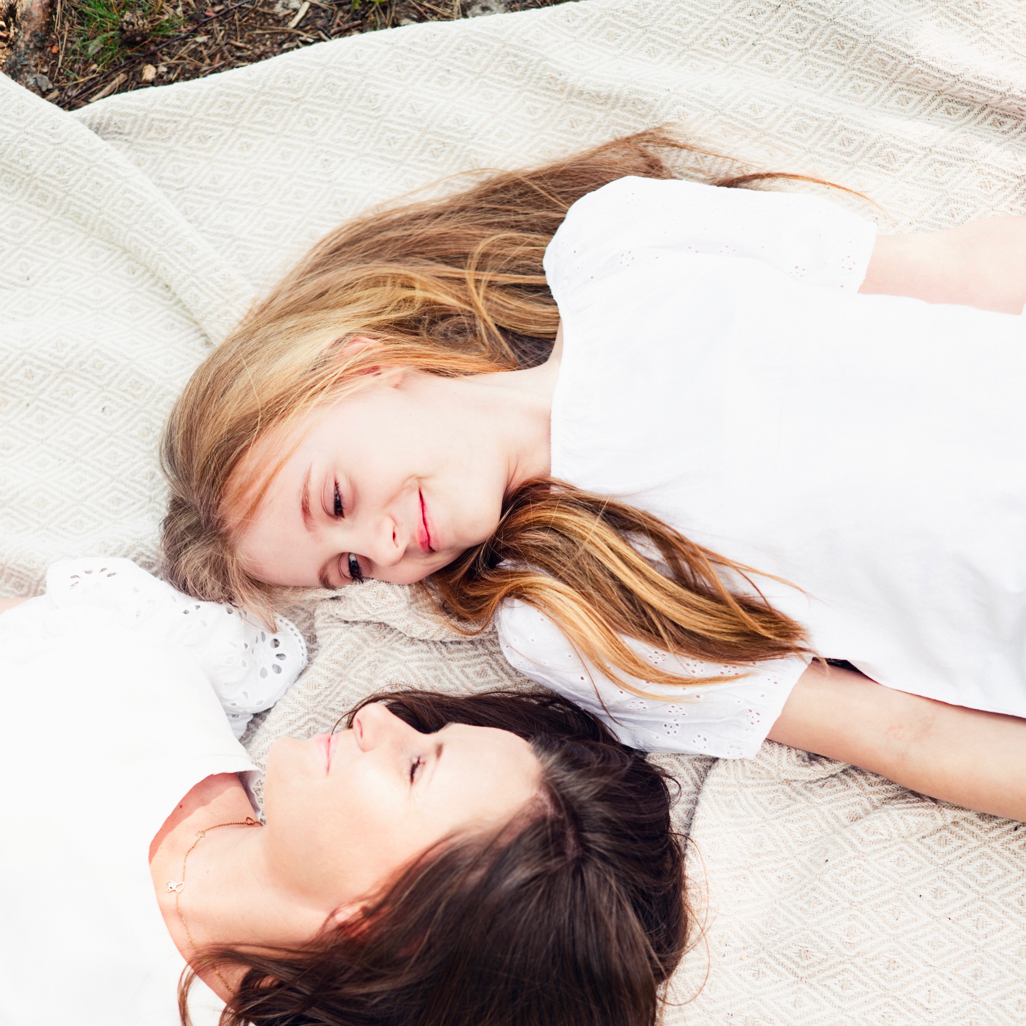 Mama i córka leżą na wzorzystym kocu w plenerze, obie ubrane w białe bluzki, ujęcie z góry.