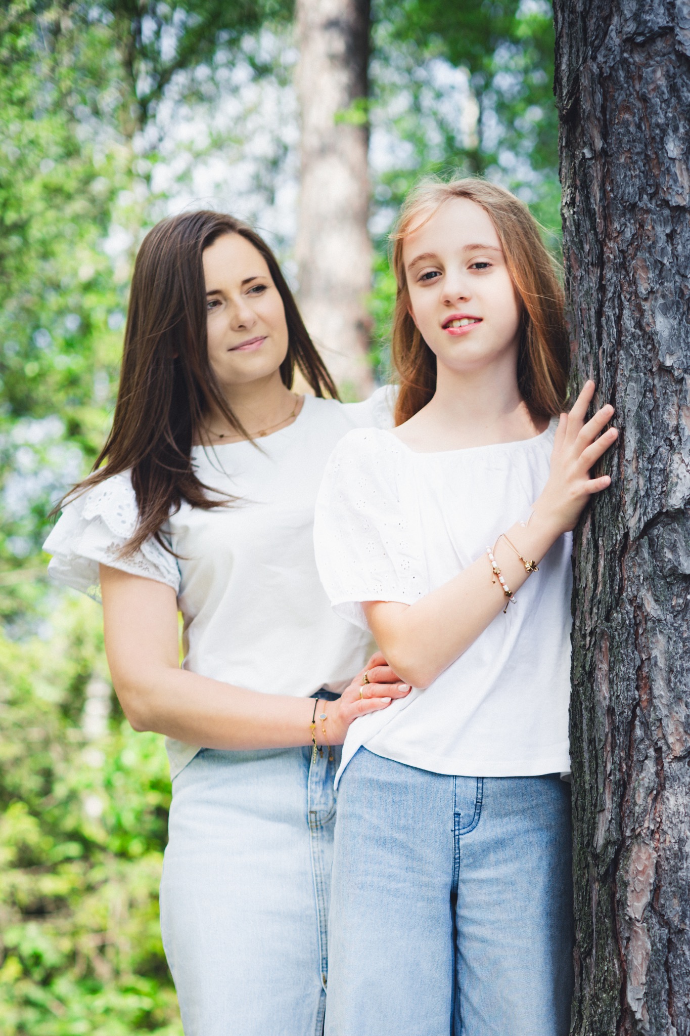 Portret matki spoglądającej z uśmiechem na córkę opierającą się o korę drzewa w leśnej scenerii, obie ubrane w białe bluzki i jasne jeansy.