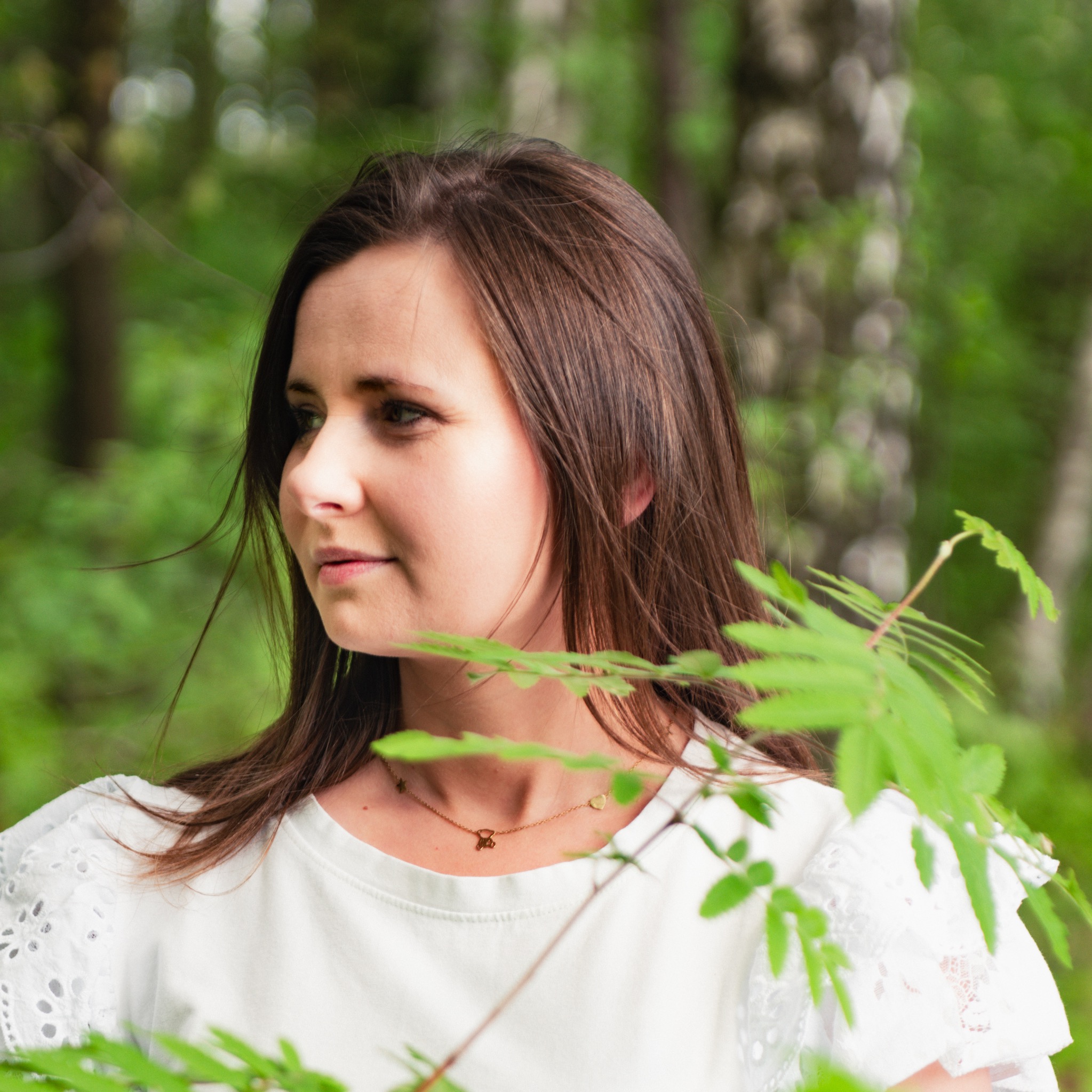 Portret kobiety z brązowymi włosami, ubrana w białą bluzkę z koronkowymi rękawami, pozująca w lesie z zielonymi liśćmi na pierwszym planie i delikatnym naszyjnikiem z motywami gwiazdki, serca i misia.