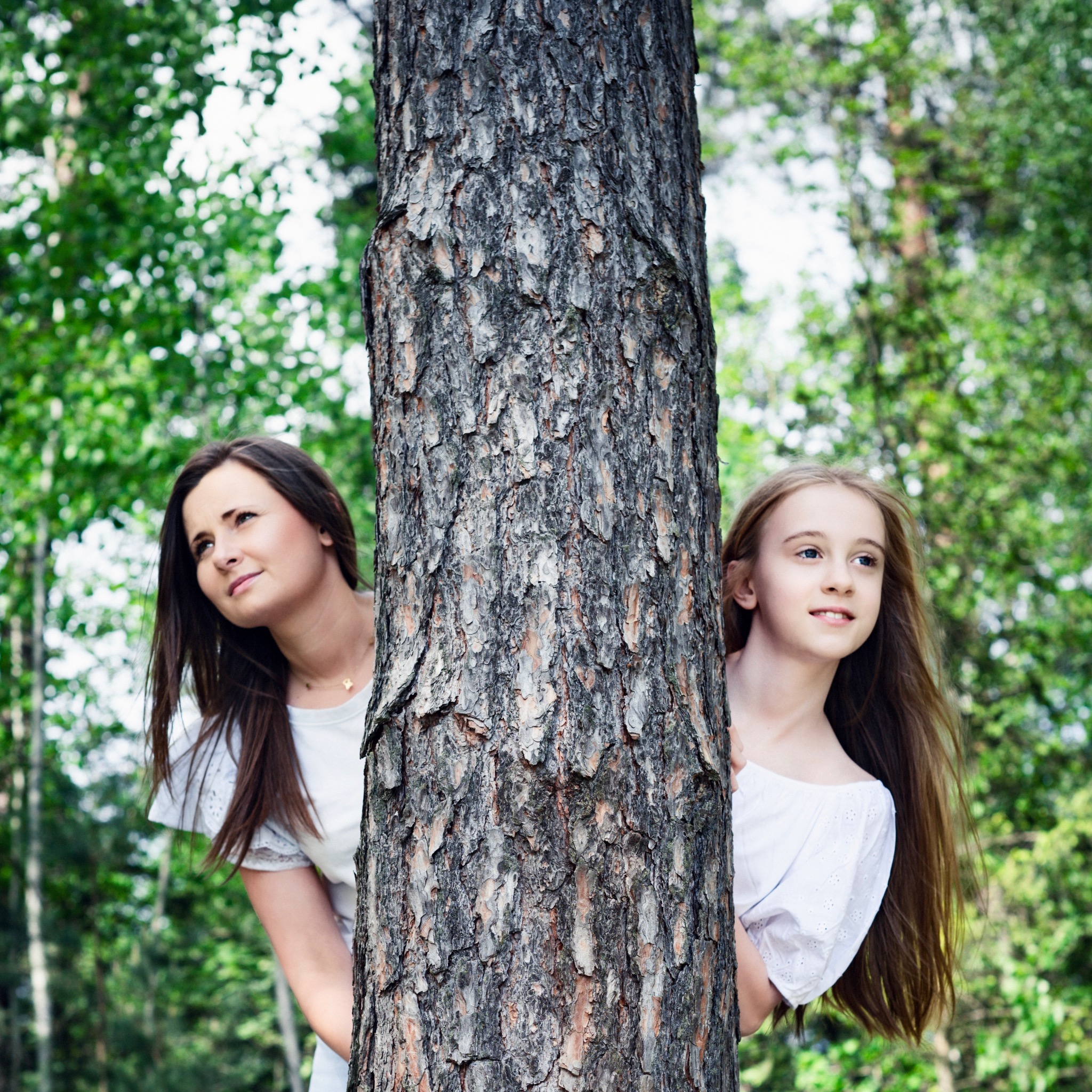 Matka z córką chowają się za pniem drzewa w lesie, obie ubrane w białe bluzki z koronkowymi detalami, patrzą w tym samym kierunku.