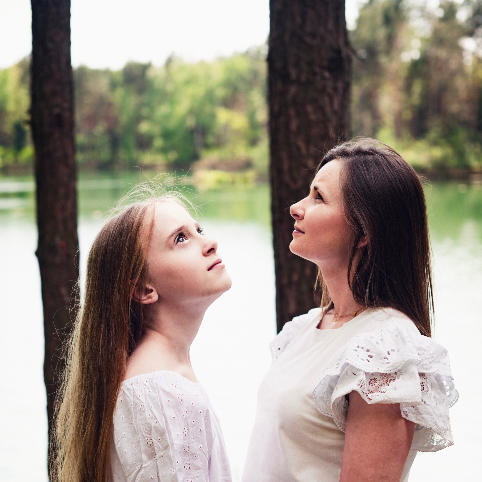 Matka z córką w białych bluzkach z koronkowymi detalami spoglądają w górę na tle drzew i jeziora, uchwycone w naturalnym świetle.