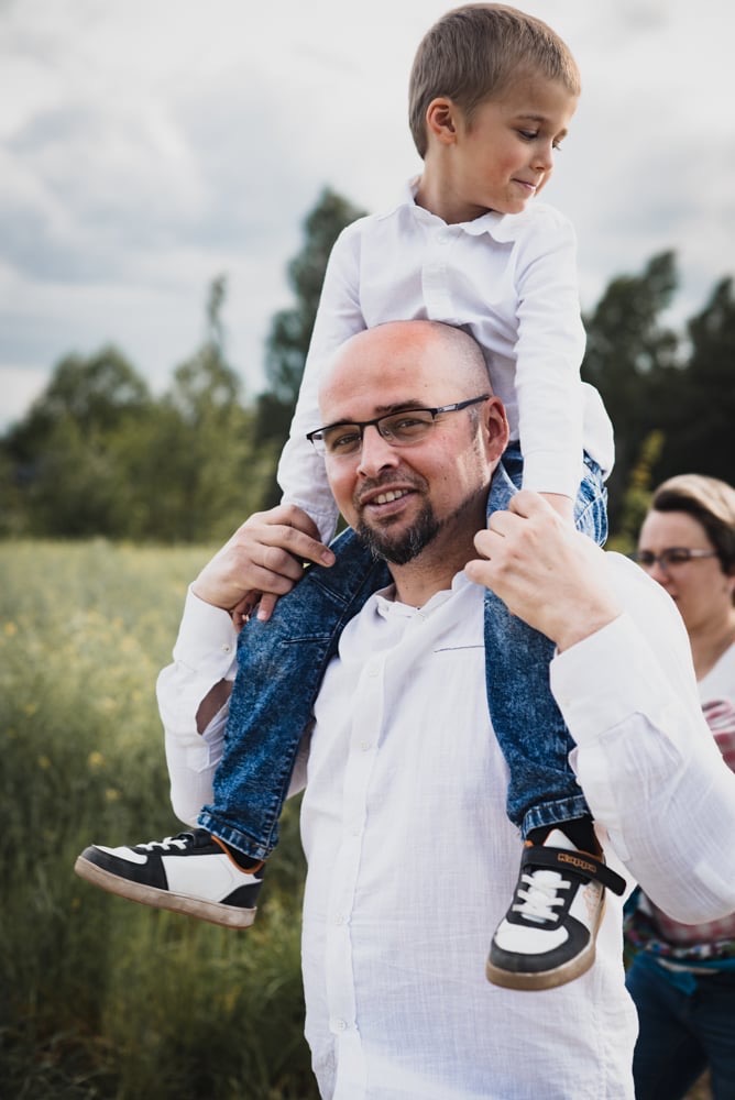 Uśmiechnięty ojciec w okularach niesie syna na barana w plenerze, w tle zieleń i fragment sylwetki kobiety.