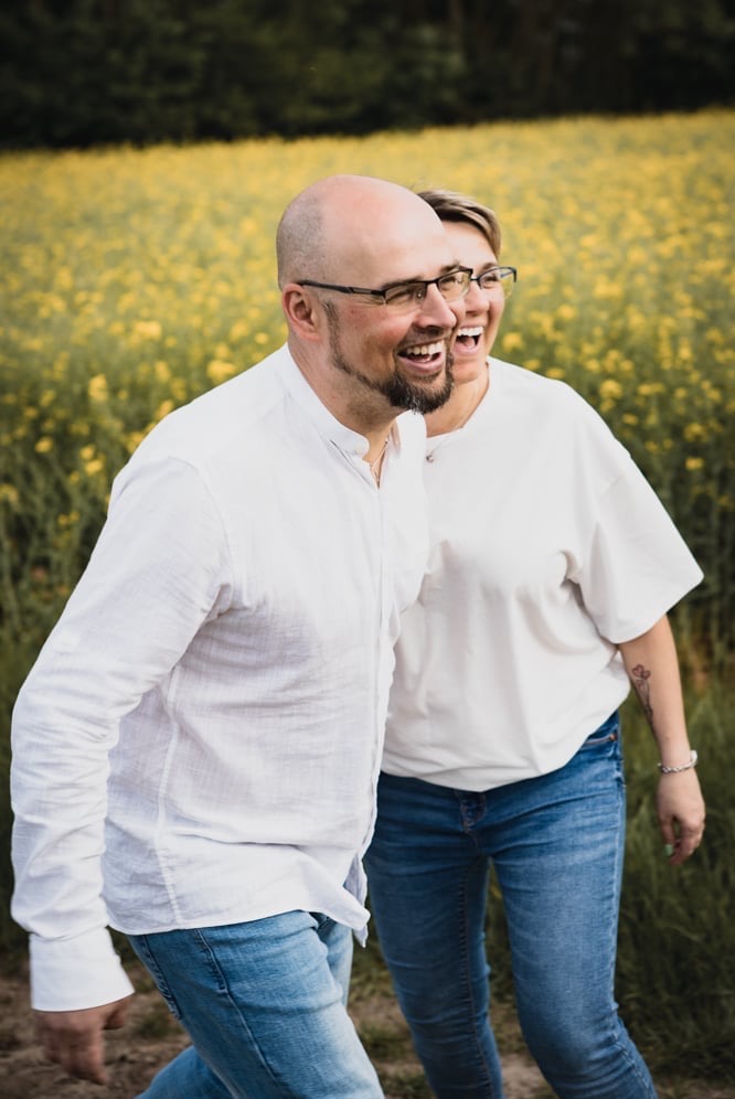 Uśmiechnięta para, mężczyzna z brodą i kobieta w okularach, spacerują po polu rzepaku, ubrani w białe koszule i dżinsy.
