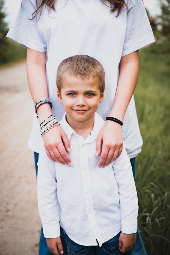 Portret uśmiechniętego chłopca w białej koszuli, z rękami starszej osoby na ramionach, w tle zielona trawa i ścieżka.