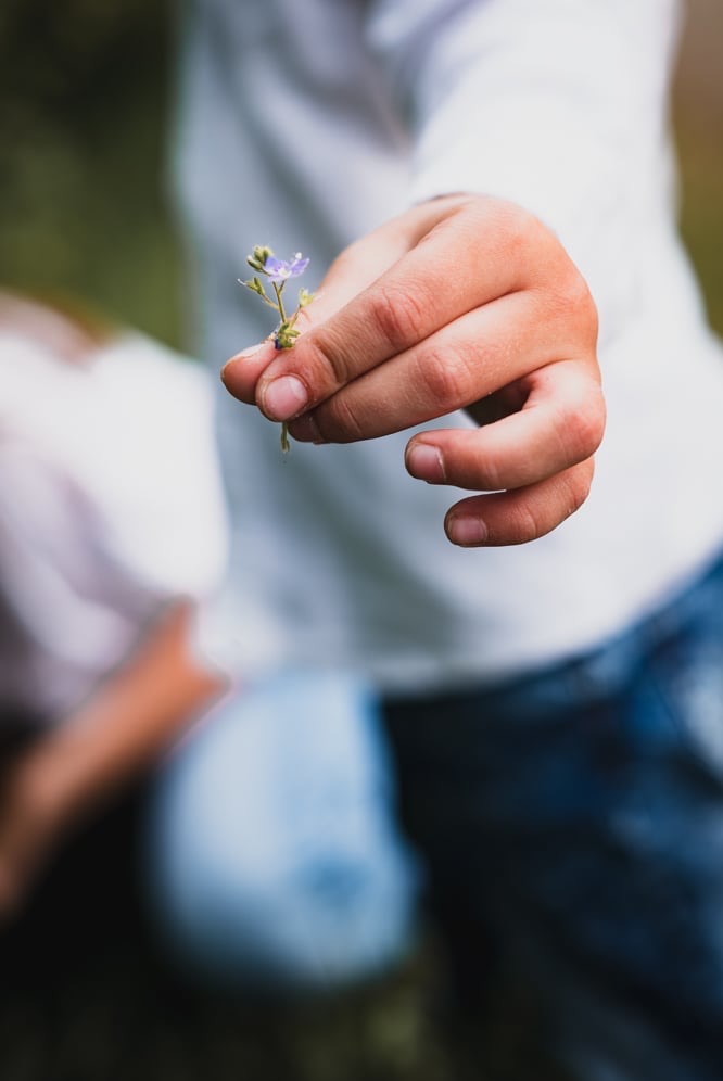 Dłoń dziecka trzymająca mały, fioletowy kwiatek na tle rozmytej postaci w białej koszuli i jeansach.