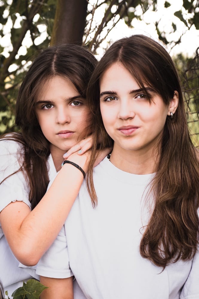 Portret dwóch młodych brunetek w białych koszulkach na tle zieleni, jedna opiera rękę na ramieniu drugiej, patrząc w obiektyw.