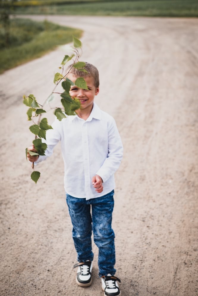 Uśmiechnięty chłopiec w białej koszuli i dżinsach, stojący na polnej drodze, trzymający gałązkę z zielonymi liśćmi, częściowo zasłaniającą twarz.