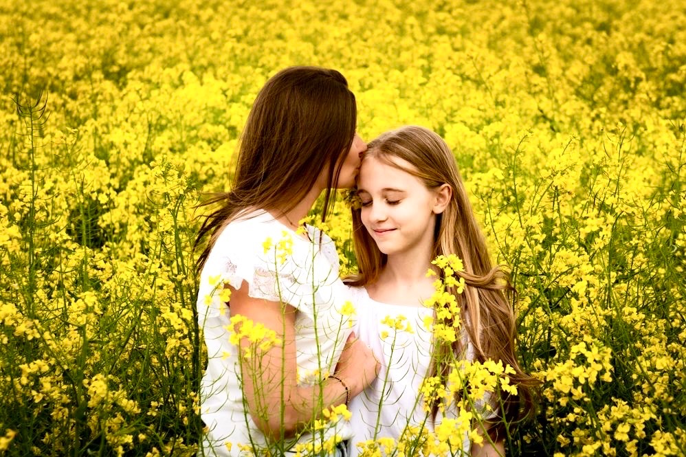 Kobieta całuje dziewczynkę w czoło na tle kwitnącego, żółtego pola rzepaku, obie ubrane na biało.