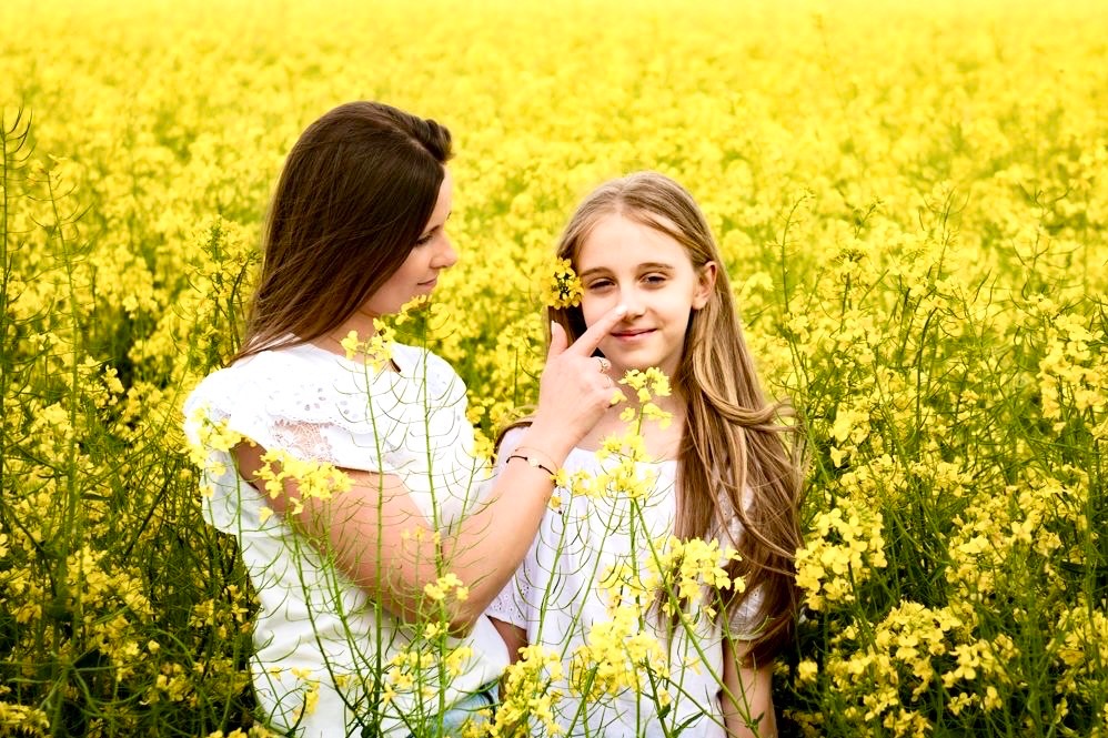 Matka delikatnie dotyka palcem nosa córki w polu kwitnącego rzepaku, obie ubrane w białe bluzki, uśmiech na twarzy dziewczynki.