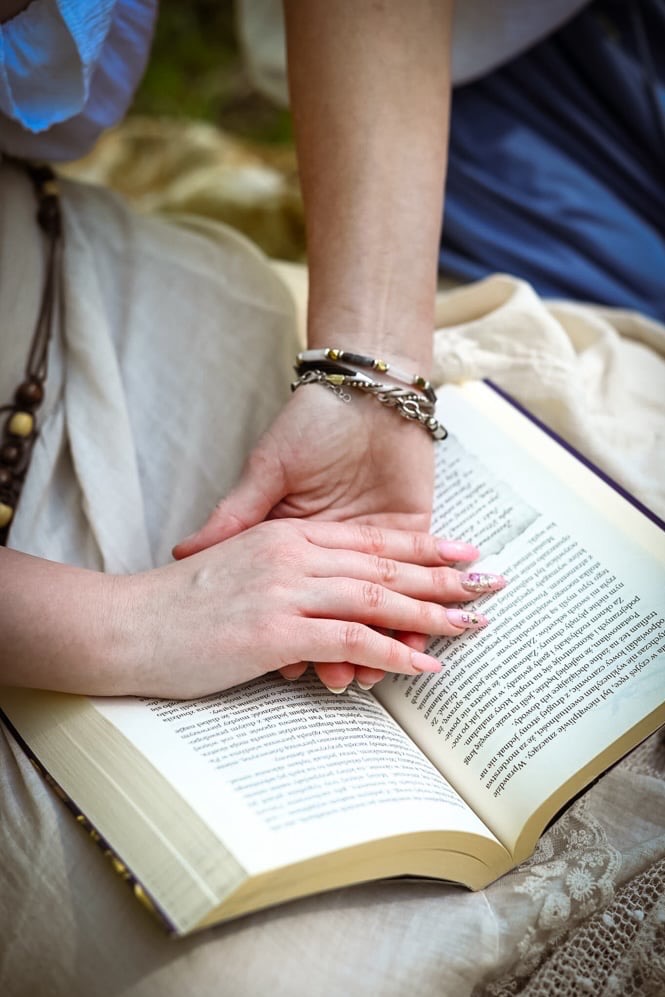Kobieta w zwiewnej sukience z drewnianym naszyjnikiem, czytająca książkę na beżowej, koronkowej tkaninie, z dłońmi ozdobionymi delikatnym manicure i bransoletkami.