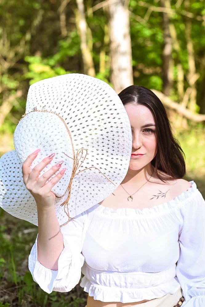 Portret kobiety z białym kapeluszem zakrywającym część twarzy, w tle rozmyta zieleń drzew.