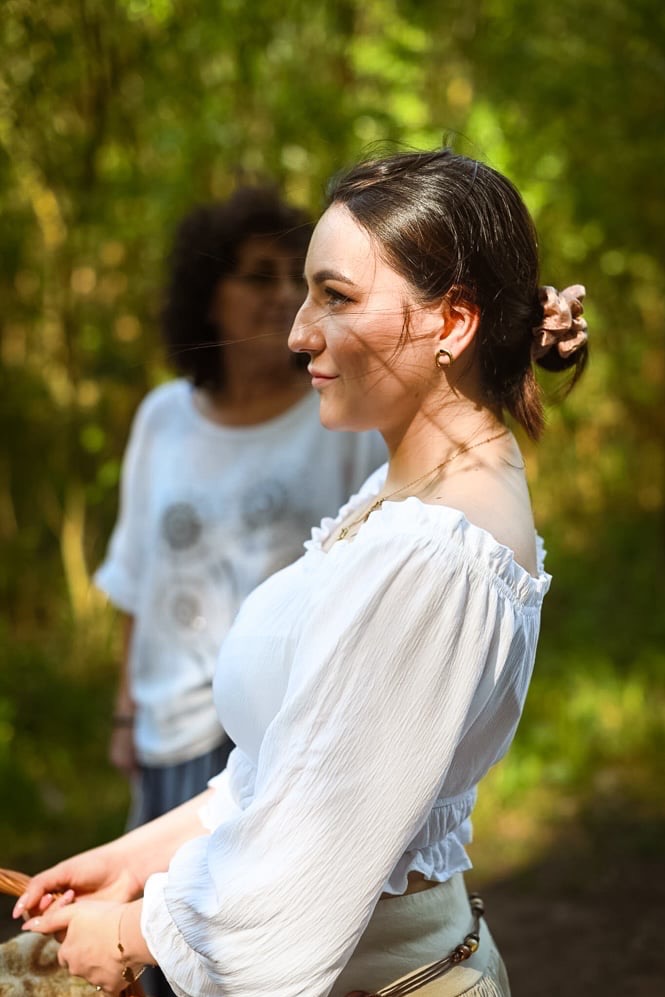 Portret profilowy młodej kobiety w białej bluzce z odkrytymi ramionami, z włosami upiętymi w kok z ozdobną gumką, na tle zamazanego, zielonego tła z drugą osobą w tle.