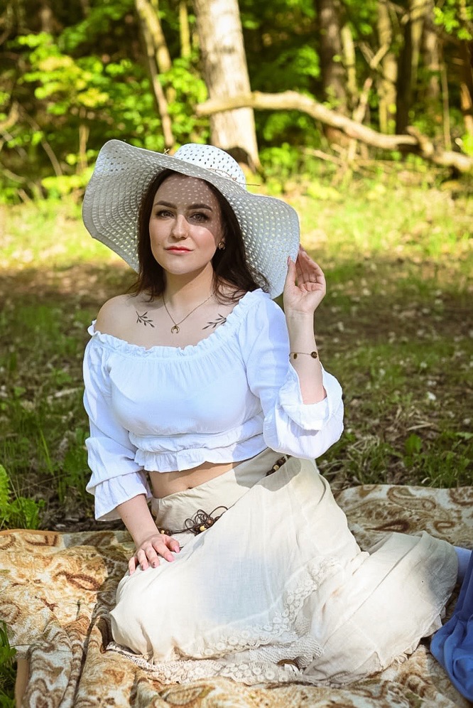 Portret młodej kobiety w białej bluzce hiszpance i kapeluszu z szerokim rondem, siedzącej na kocu w plenerze.