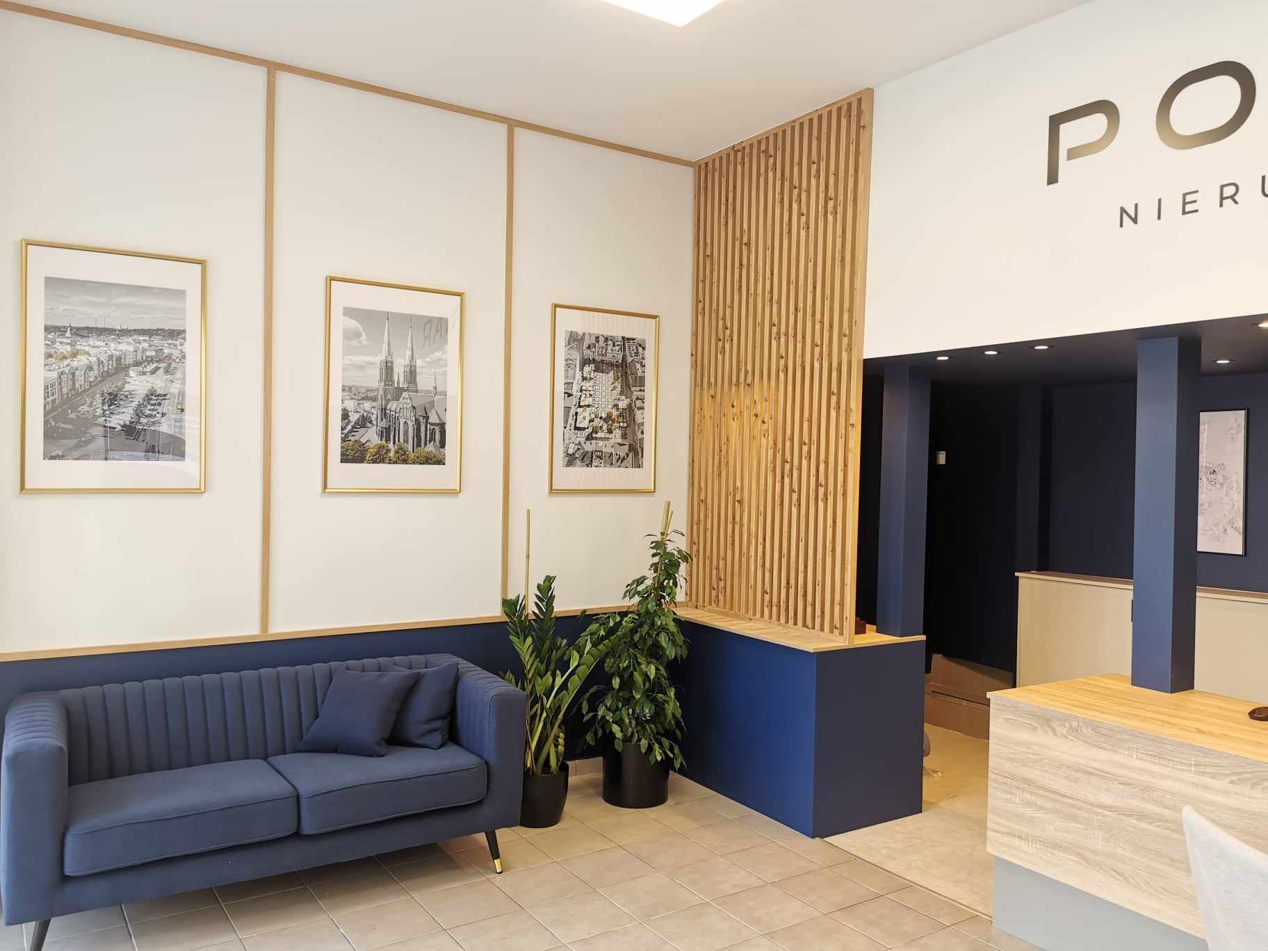 Wnętrze biura nieruchomości z niebieską sofą, roślinami doniczkowymi i grafikami w złotych ramach na ścianach, widoczny fragment napisu 'PO NIERU'.