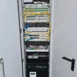 Otwarta szafa rack z zainstalowanymi urządzeniami telekomunikacyjnymi, widoczne kable światłowodowe, patch panele i routery, starannie ułożone okablowanie w kolorach ż&oacute;łtym i niebieskim.