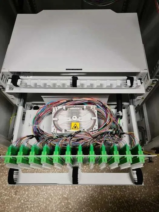 Rozdzielnica światłowodowa z licznymi kolorowymi kablami i zielonymi złączkami, widoczny znak ostrzegawczy lasera.