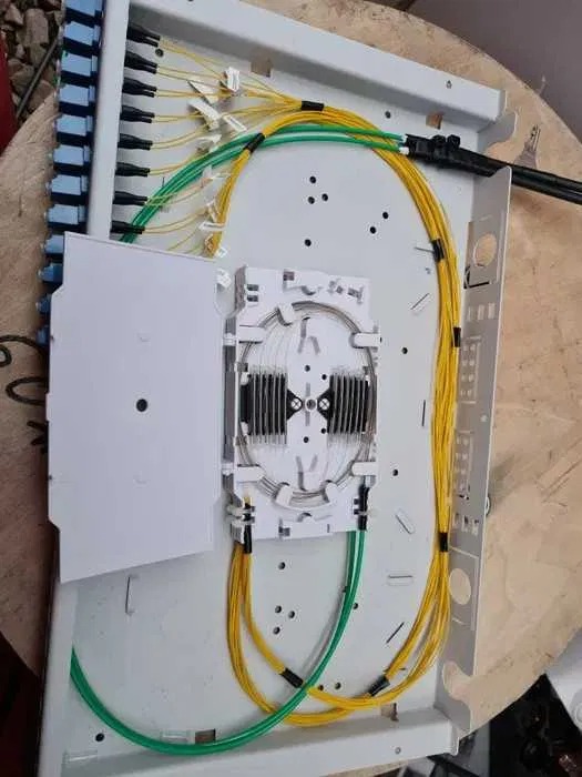 Otwarta skrzynka światłowodowa z widocznymi złączami, patchcordami w kolorach żółtym i zielonym oraz elementami mocującymi, umieszczona na drewnianym podłożu.