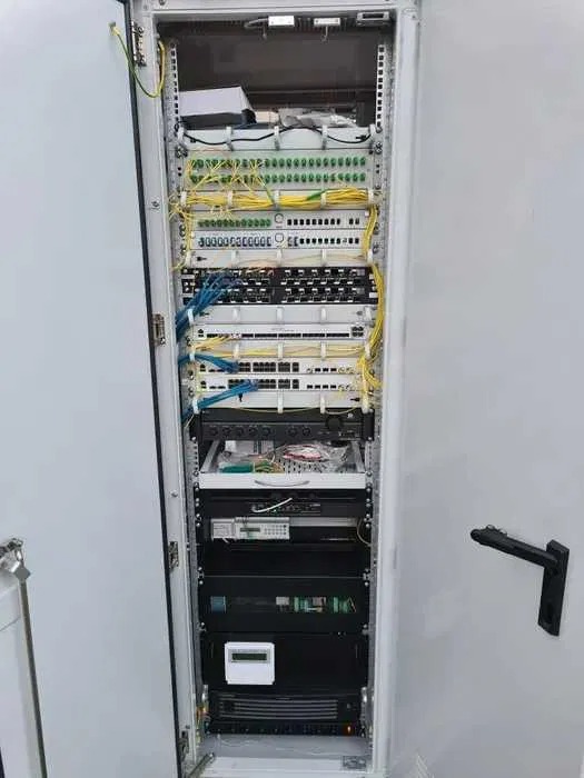 Otwarta szafa rack z zainstalowanymi urządzeniami telekomunikacyjnymi, widoczne kable światłowodowe, patch panele i routery, starannie ułożone okablowanie w kolorach żółtym i niebieskim.