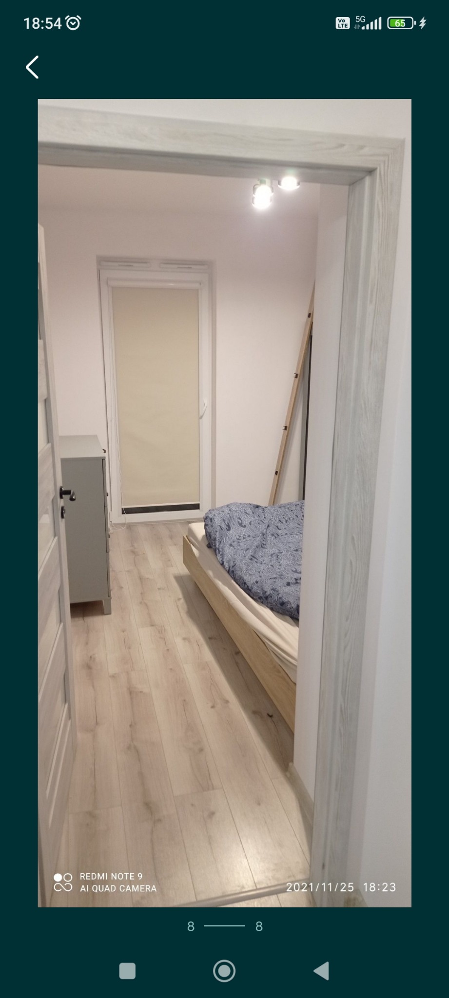 Widok na wnętrze pokoju z drewnianą podłogą i łóżkiem z niebieską pościelą, widziany zza futryny drzwi o jasnej barwie drewna, z komodą po lewej stronie i drabiną opartą o ścianę w głębi.