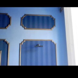 Zal - Bud - Niebieska ściana z dekoracyjnymi panelami w paski, obramowanymi złotą listwą, oświetlona górnym światłem