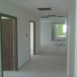 Zal - Bud - Korytarz biurowy w trakcie prac wykończeniowych, widoczne białe ściany, sufit z otworami, częściowo zamontowane drzwi i elementy instalacji.