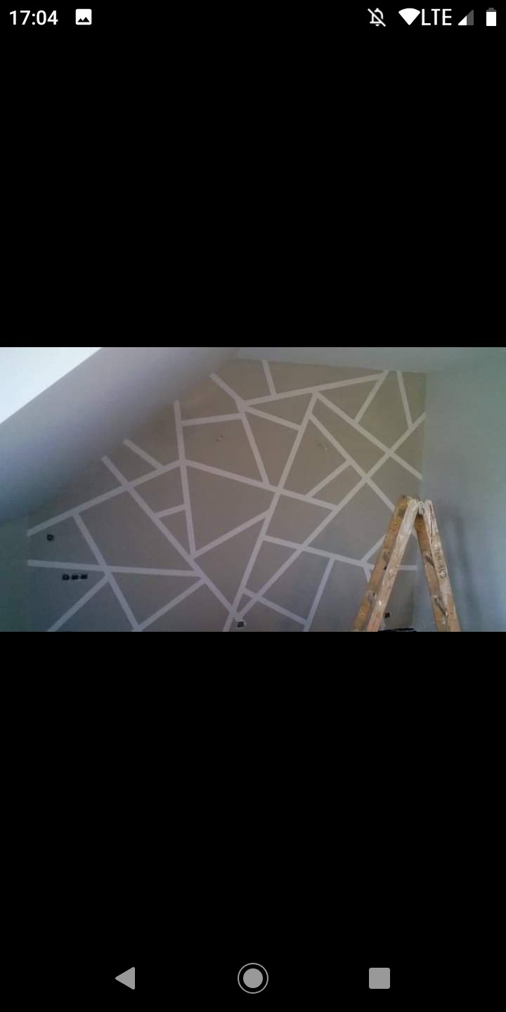 Ściana w pokoju na poddaszu przygotowana do malowania z geometrycznym wzorem wyklejonym taśmą malarską, drabina oparta o ścianę, widoczne puszki elektryczne.