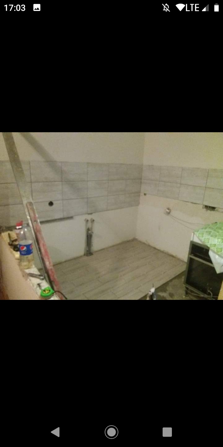 Wnętrze w trakcie remontu: częściowo wyłożone płytkami ściany, ułożona podłoga z paneli, widoczne rury i butelka napoju na parapecie.