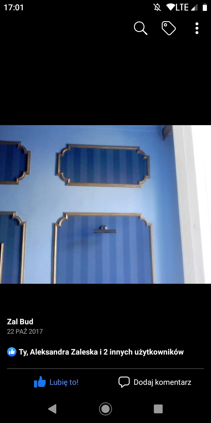 Niebieska ściana z dekoracyjnymi panelami w paski, obramowanymi złotą listwą, oświetlona górnym światłem