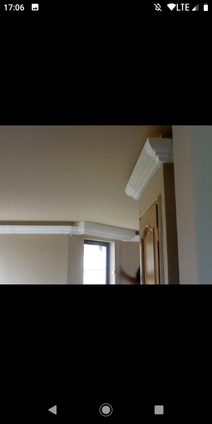 Montaż białych listew dekoracyjnych na styku ściany i sufitu w jasnym wnętrzu, widoczny fragment okna i drzwi.