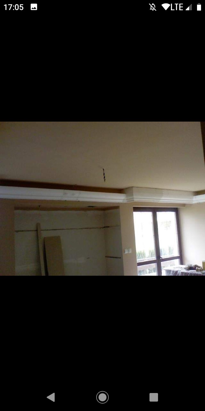 Wnętrze w trakcie prac wykończeniowych, widoczny fragment sufitu z zamontowaną sztukaterią, ściany przygotowane do malowania, okno z widokiem na zewnątrz.