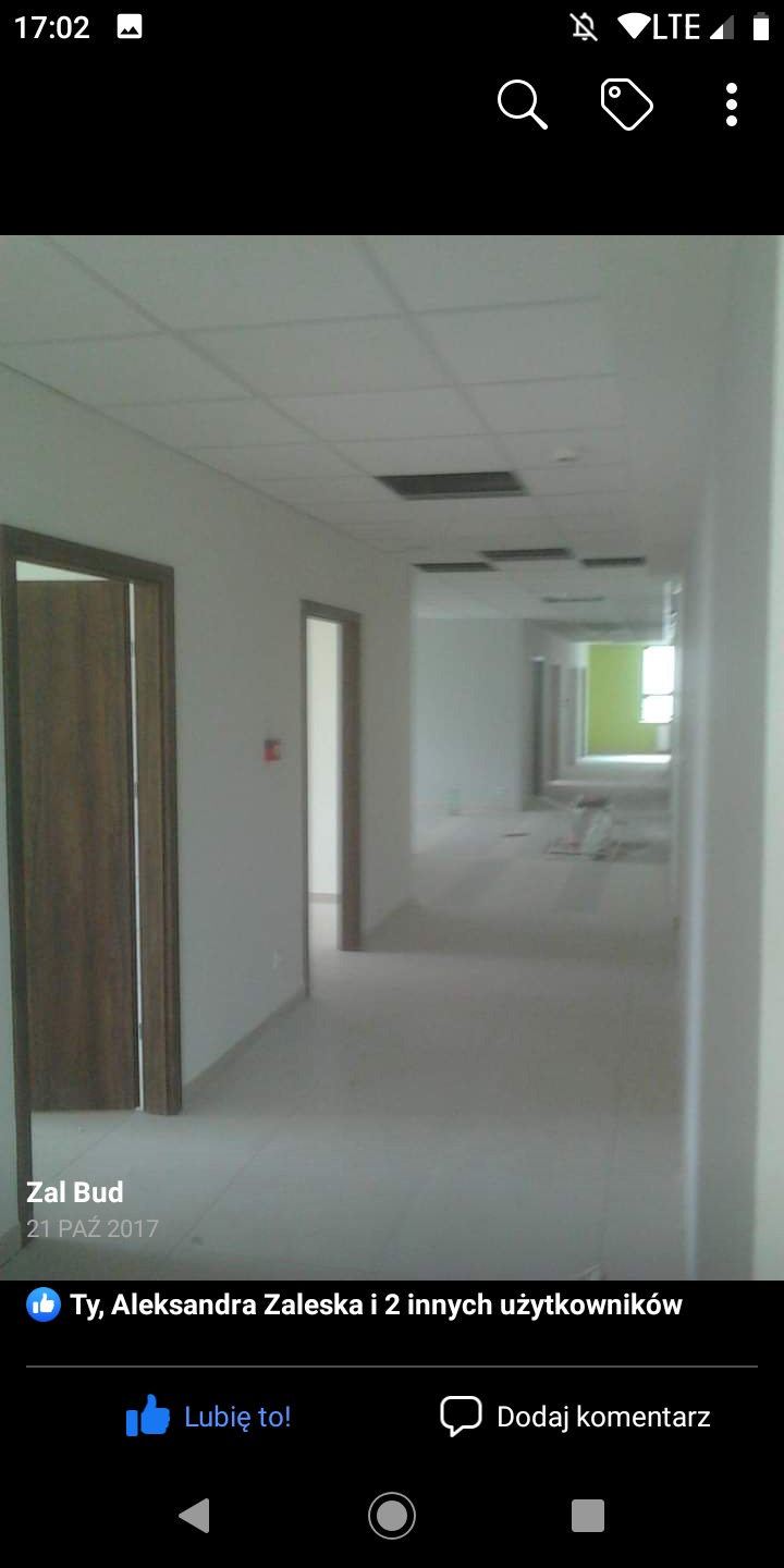 Korytarz biurowy w trakcie prac wykończeniowych, widoczne białe ściany, sufit z otworami, częściowo zamontowane drzwi i elementy instalacji.