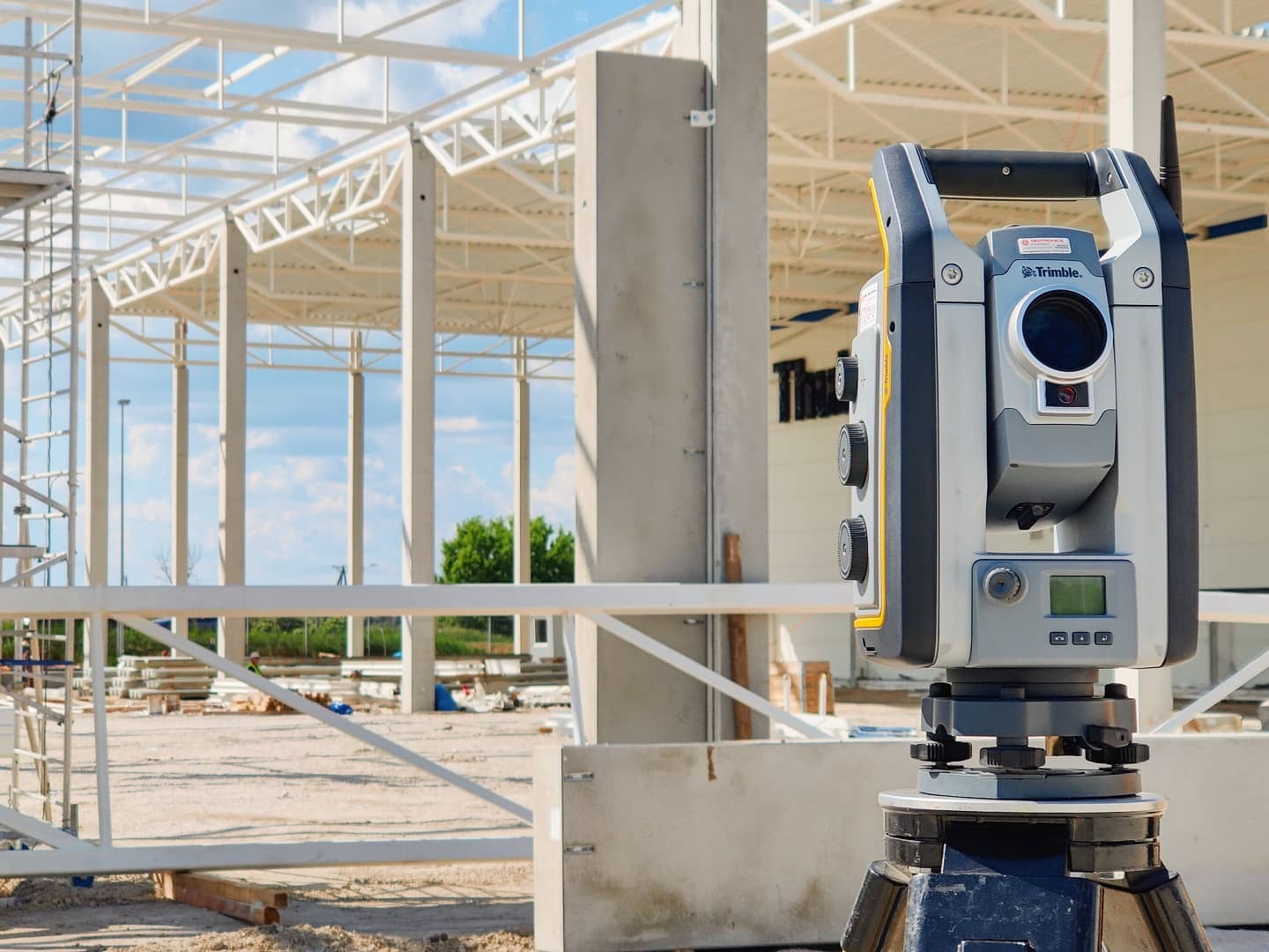 Profesjonalny teodolit Trimble na trójnogu na tle konstrukcji stalowej budynku w budowie, widoczne elementy betonowe i rusztowania.