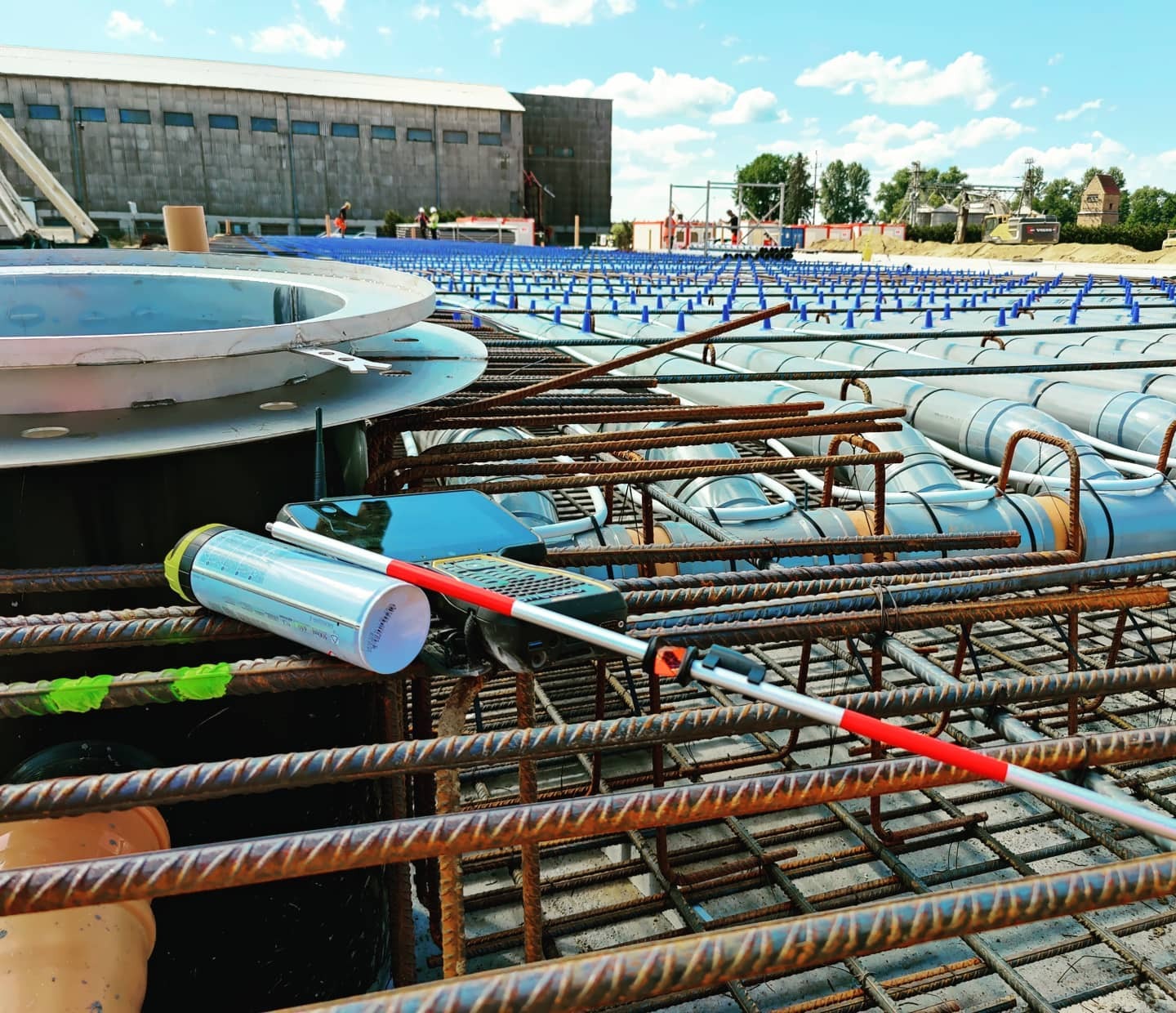 Przybory pomiarowe (tyczka geodezyjna, odbiornik GPS) leżące na zbrojeniu fundamentu, w tle widoczny budynek przemysłowy i niebo z chmurami.
