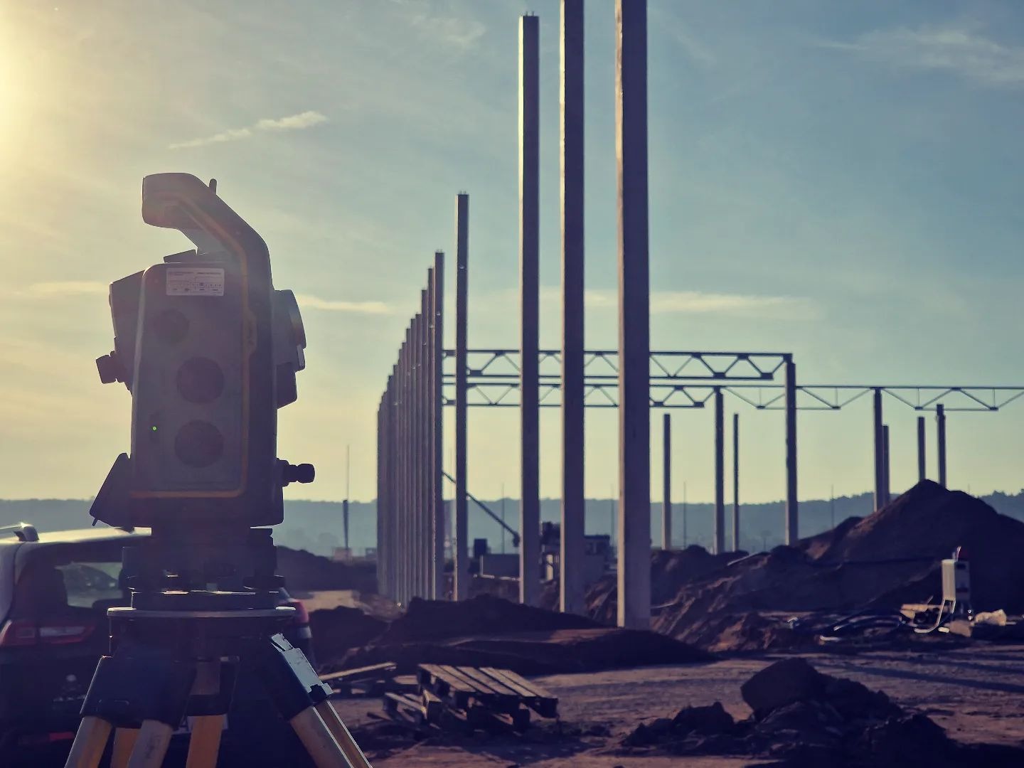 Geodezyjny teodolit na statywie ustawiony na placu budowy, w tle widoczne słupy konstrukcji hali w surowym stanie, z jasnym słońcem w górnym lewym rogu kadru.