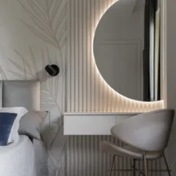 Nowoczesna sypialnia z toaletką: Okrągłe lustro z podświetleniem LED, kremowe panele ścienne z pionowymi żłobieniami, tapeta z motywem liści, szara tapicerowana rama łóżka i krzesło z beżową...