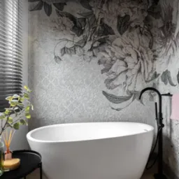 Nowoczesna biała wanna wolnostojąca z czarną armaturą na tle ściany z tapetą w motywy kwiatowe w odcieniach szarości, drewniana podłoga ułożona w jodełkę, stolik pomocniczy z akcesoriami kąpielowymi.
