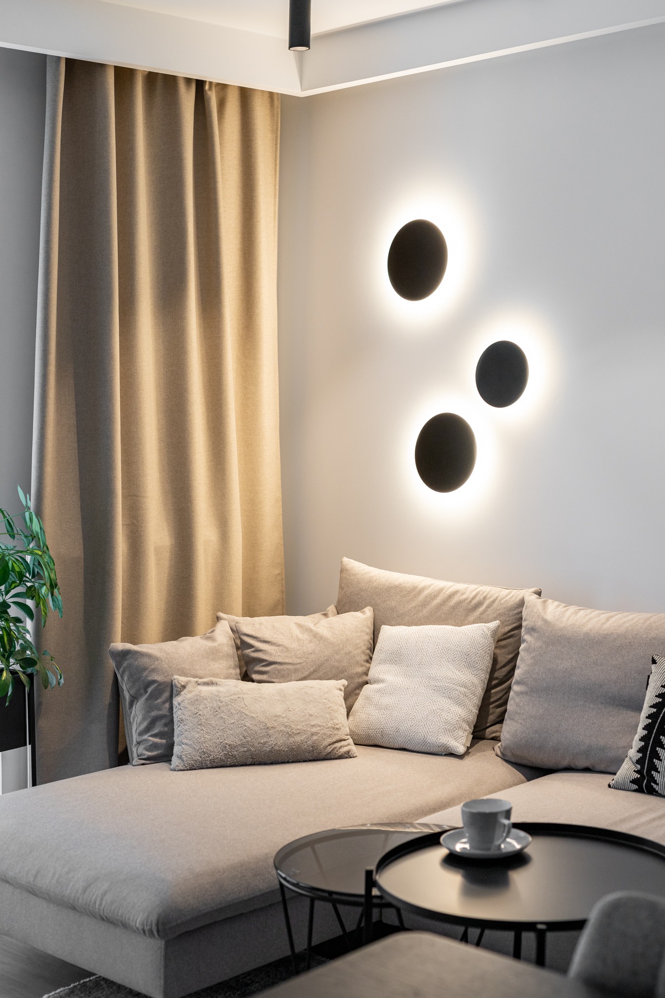 Beżowa sofa z poduszkami w salonie, okrągłe stoliki kawowe z filiżanką, dekoracyjne okrągłe lampy ścienne, zasłony i roślina w doniczce.