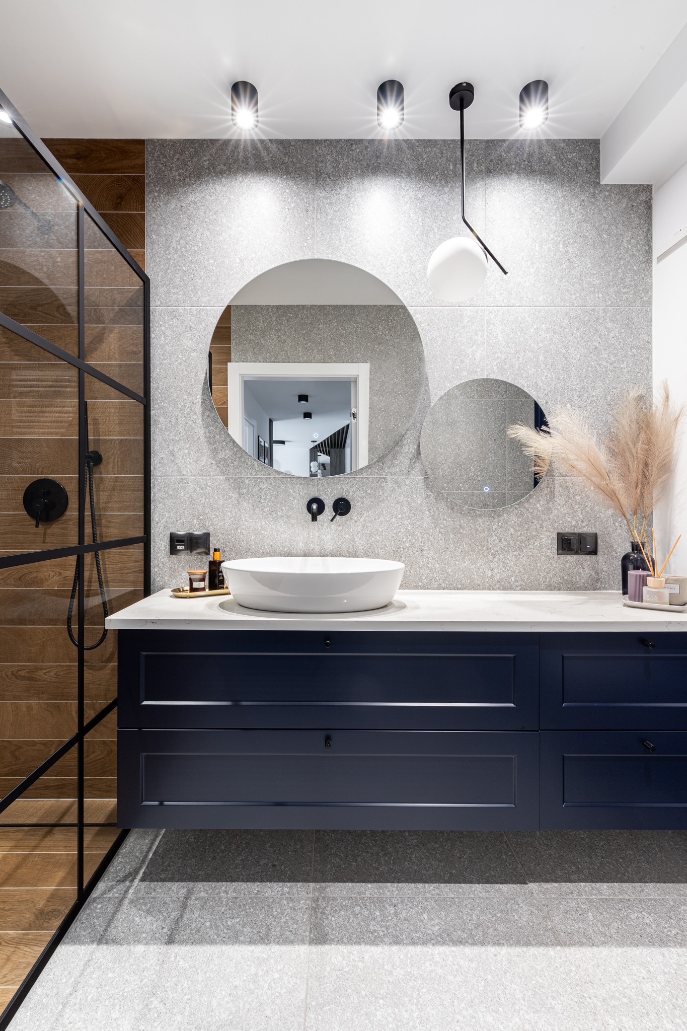 Nowoczesna łazienka z szarą mozaiką na ścianach, ciemnoniebieską szafką pod umywalką, okrągłymi lustrami i kabiną prysznicową ze szklaną ścianą i czarnymi profilami.
