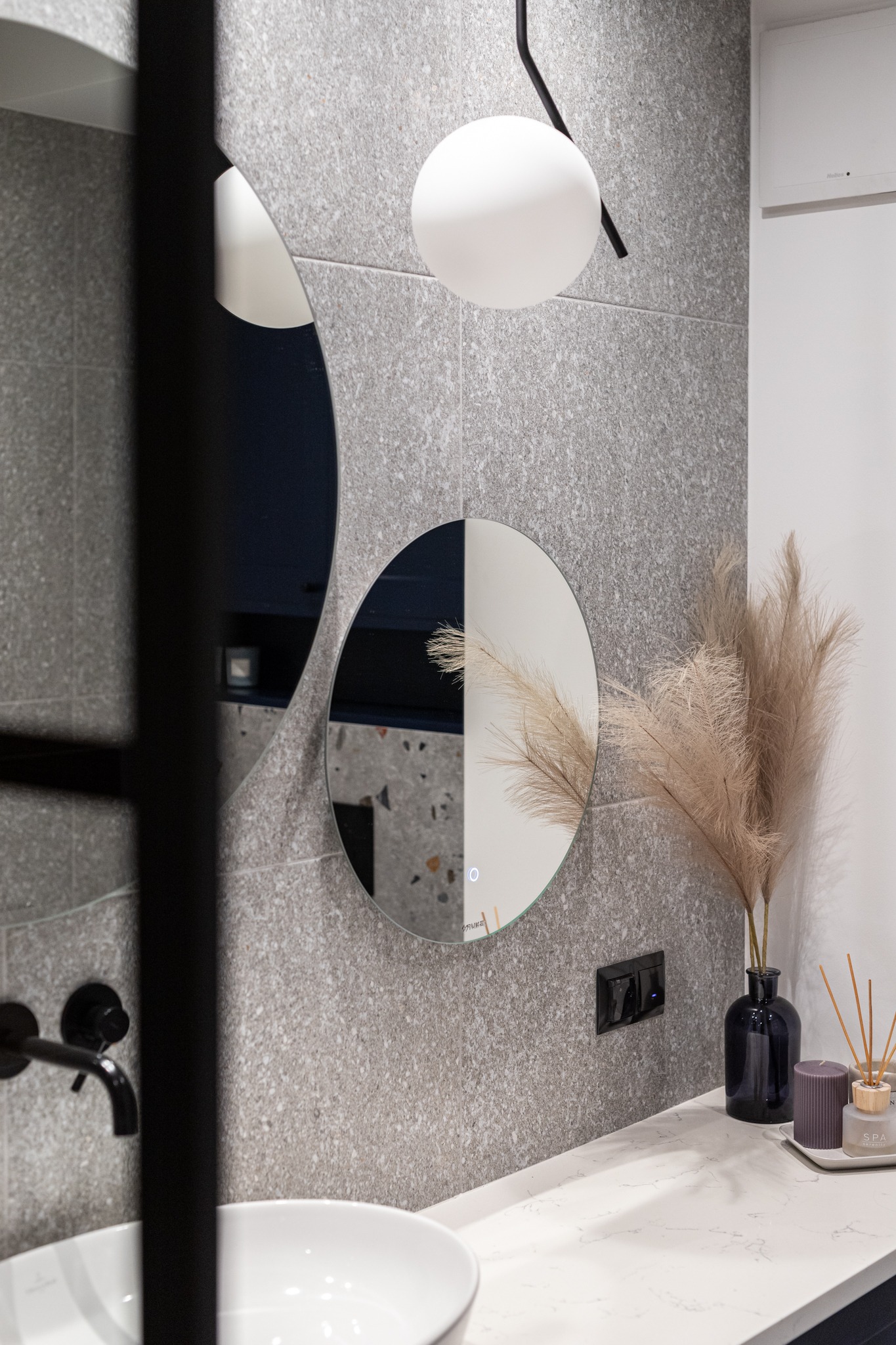 Nowoczesna łazienka z okrągłymi lustrami z podświetleniem LED, szarą płytką imitującą kamień, czarną armaturą i dekoracyjnym wazonem z suszonymi trawami pampasowymi, uzupełniona kulistą lampą wiszącą.