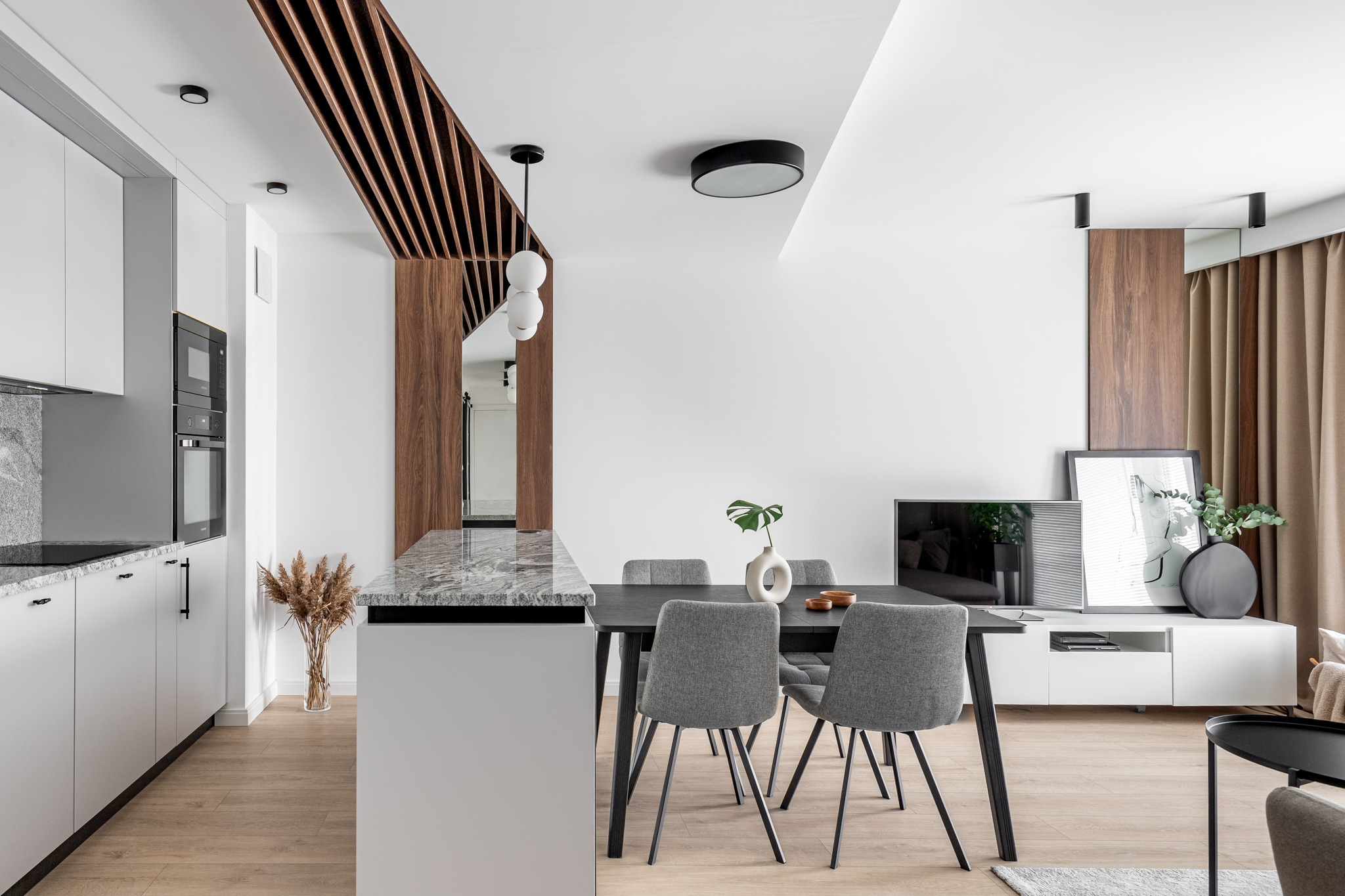 Nowoczesne wnętrze mieszkania z otwartą kuchnią i jadalnią, jasne ściany, drewniane akcenty dekoracyjne na suficie i ścianie, szary stół jadalny z krzesłami w odcieniach szarości, minimalistyczne...