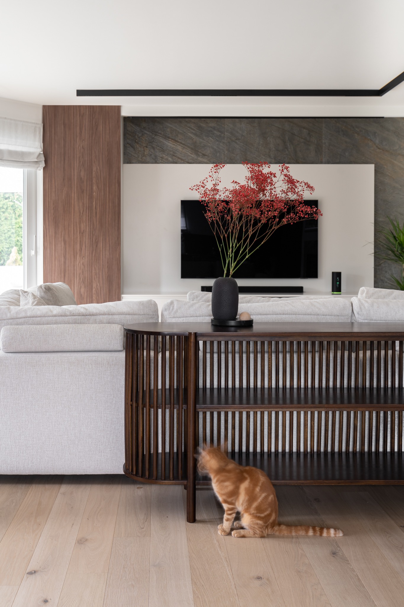 Jasny salon z drewnianą konsolą, na której stoi czarny wazon z czerwonymi kwiatami. W tle telewizor na białej ścianie z elementami z szarego kamienia. Na pierwszym planie rudy kot siedzący...
