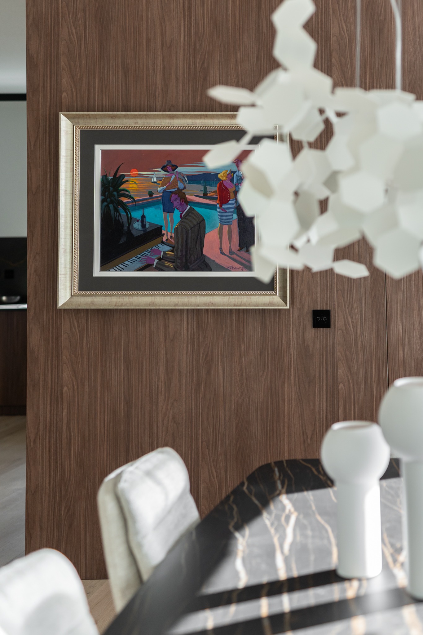 Fragment eleganckiego wnętrza z drewnianą ścianą, na której wisi obraz w złotej ramie przedstawiający scenę przy basenie z pianistą; widoczny fragment marmurowego stołu z białymi wazonami i białe...