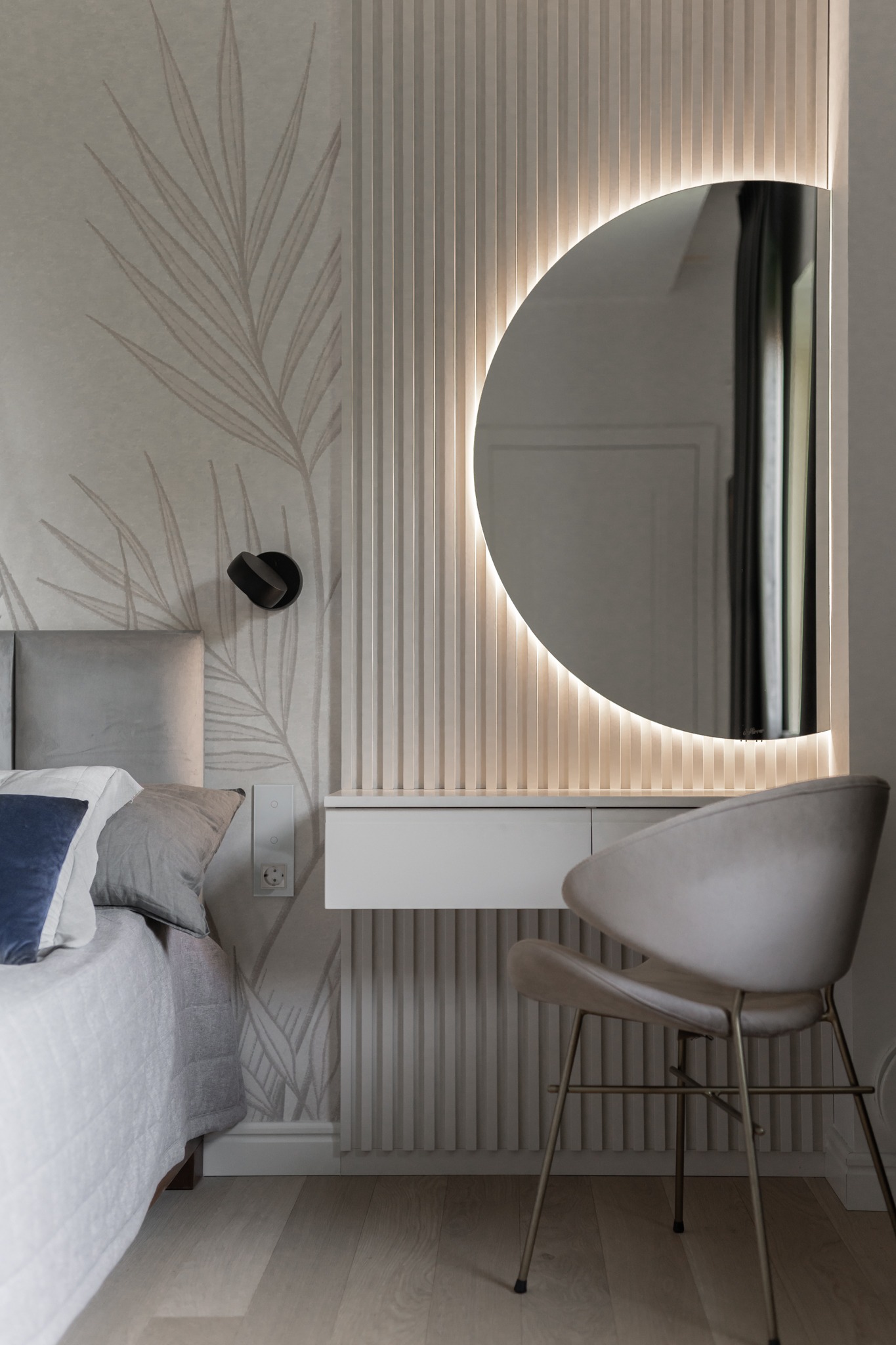 Nowoczesna sypialnia z toaletką: Okrągłe lustro z podświetleniem LED, kremowe panele ścienne z pionowymi żłobieniami, tapeta z motywem liści, szara tapicerowana rama łóżka i krzesło z beżową...