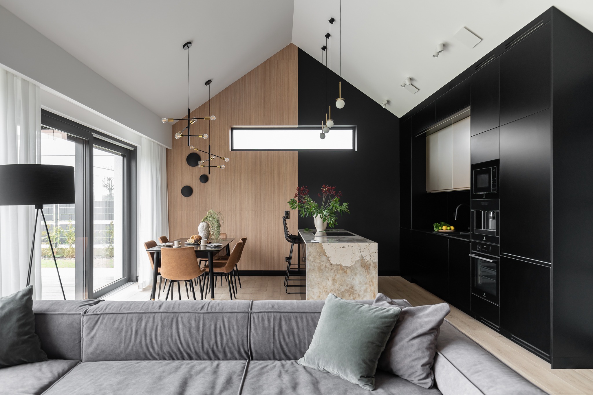 Nowoczesne wnętrze domu z otwartą kuchnią, jadalnią i salonem, charakteryzujące się kontrastowym połączeniem drewnianej i czarnej ściany, wyspą kuchenną z blatem imitującym marmur oraz szarą sofą...