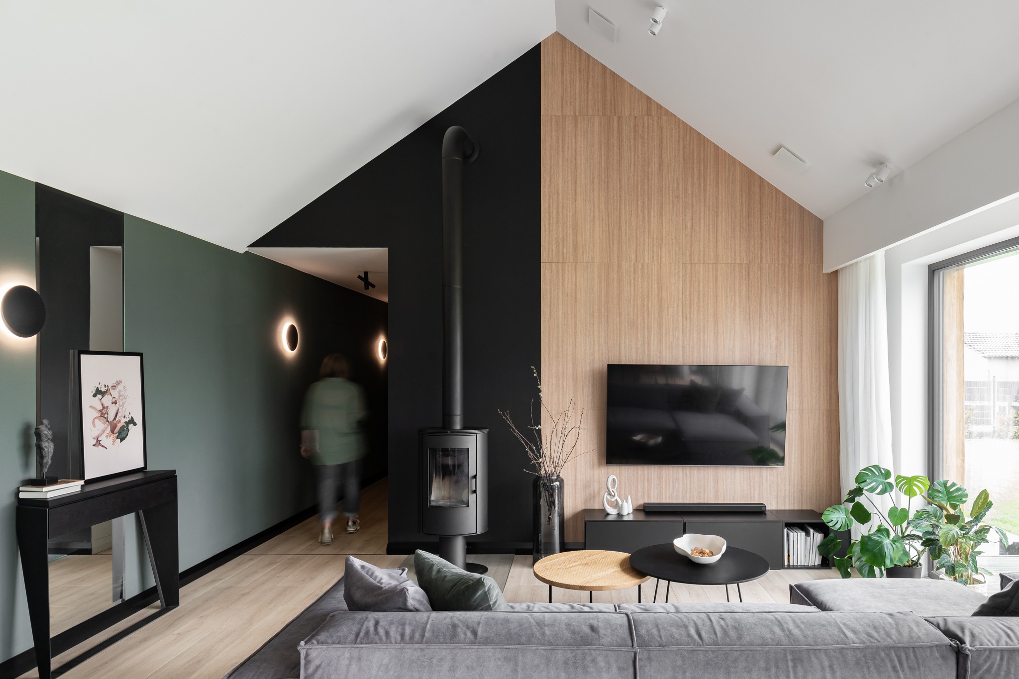 Nowoczesny salon z kominkiem, drewnianą ścianą telewizyjną i szarą sofą, oświetlony lampami ściennymi, z widoczną osobą w tle przechodzącą przez korytarz.