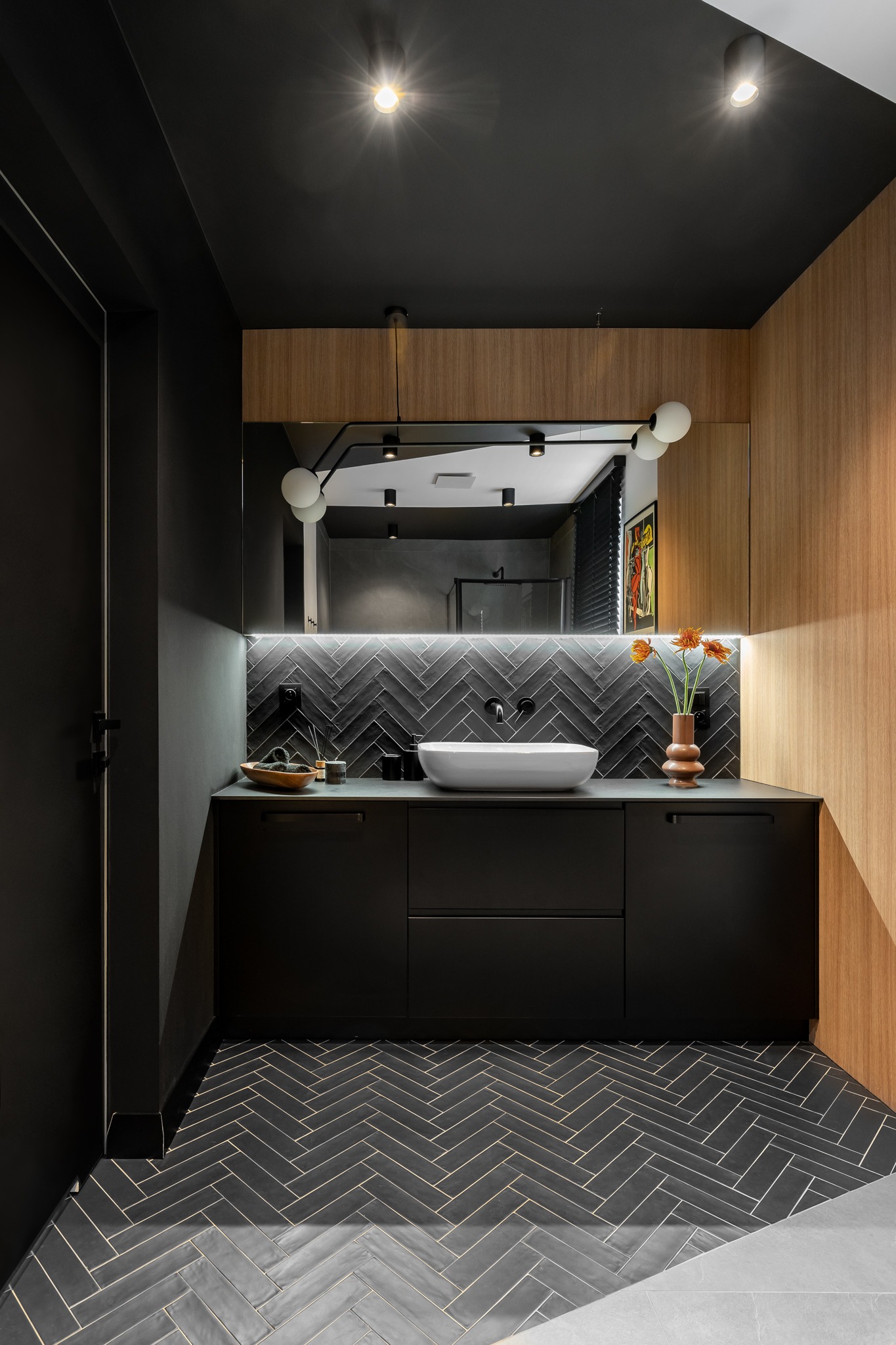 Nowoczesna łazienka z czarnymi płytkami ułożonymi w jodełkę na podłodze, czarnymi szafkami pod umywalką nablatową, drewnianą okładziną ściany i oświetleniem punktowym, akcentowana pomarańczowymi...