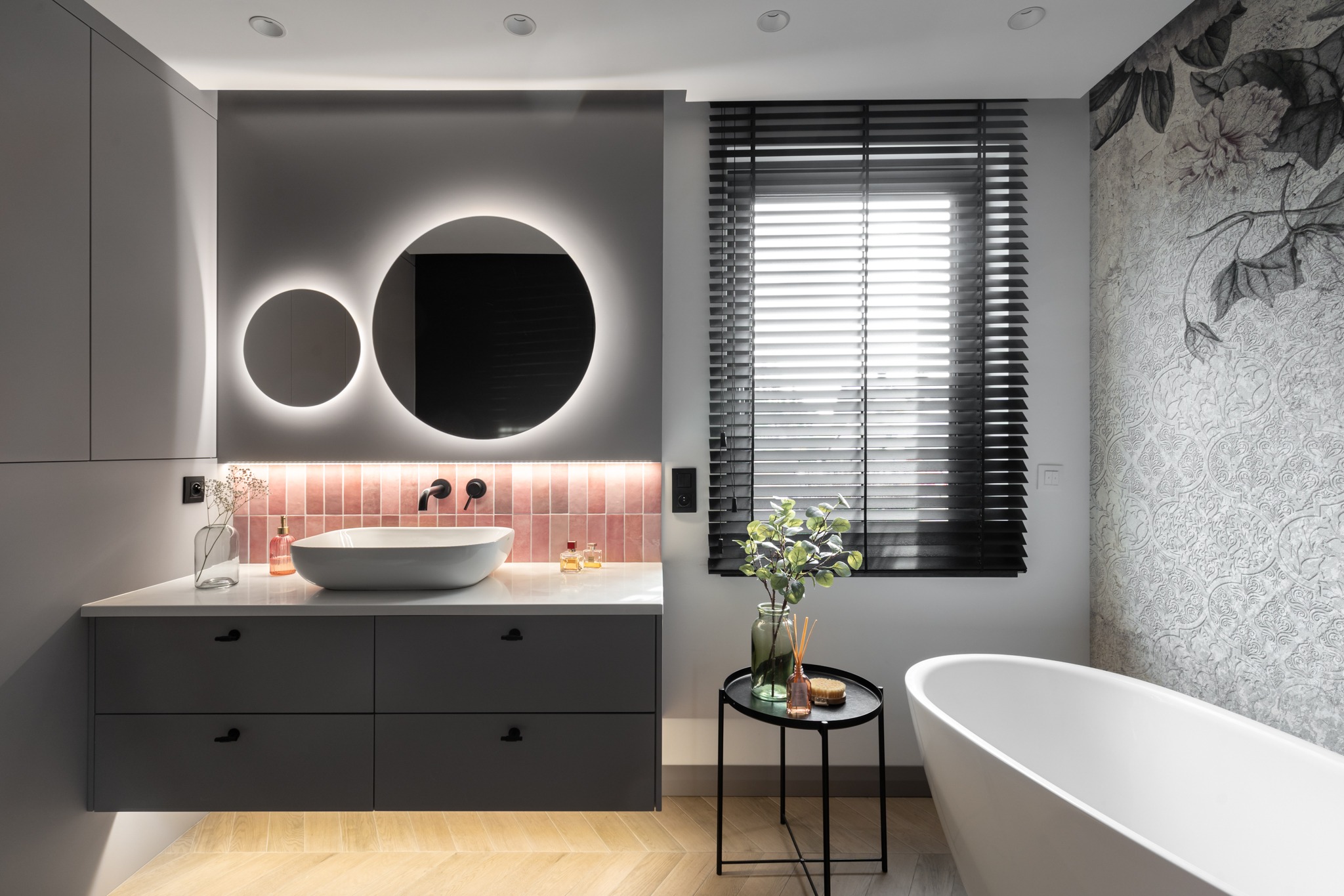 Nowoczesna łazienka z okrągłym lustrem podświetlanym LED, umywalką nablatową, szarą szafką z czarnymi uchwytami, różowymi płytkami, drewnianą podłogą ułożoną w jodełkę i wolnostojącą wanną na tle...