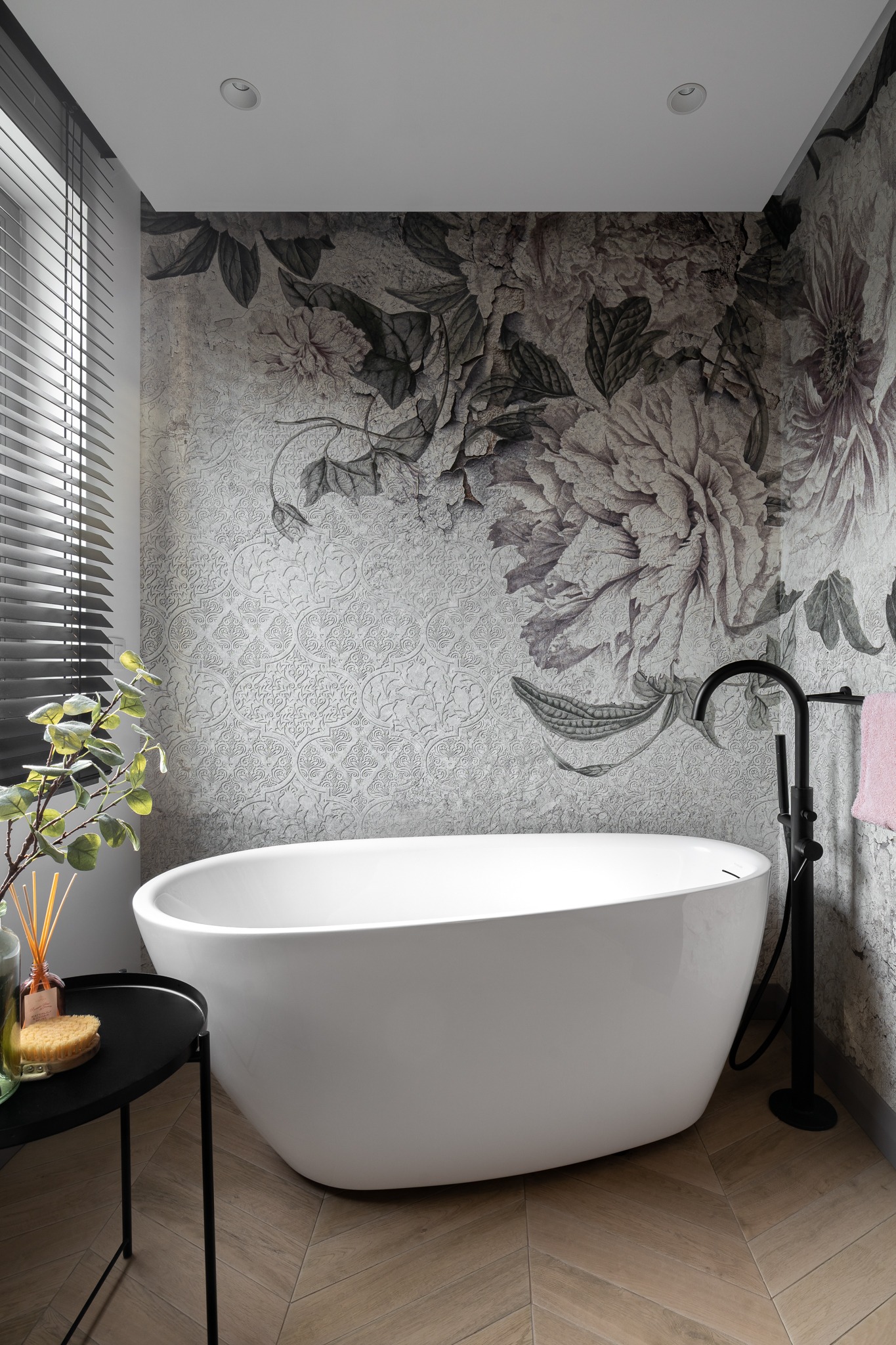 Nowoczesna biała wanna wolnostojąca z czarną armaturą na tle ściany z tapetą w motywy kwiatowe w odcieniach szarości, drewniana podłoga ułożona w jodełkę, stolik pomocniczy z akcesoriami kąpielowymi.